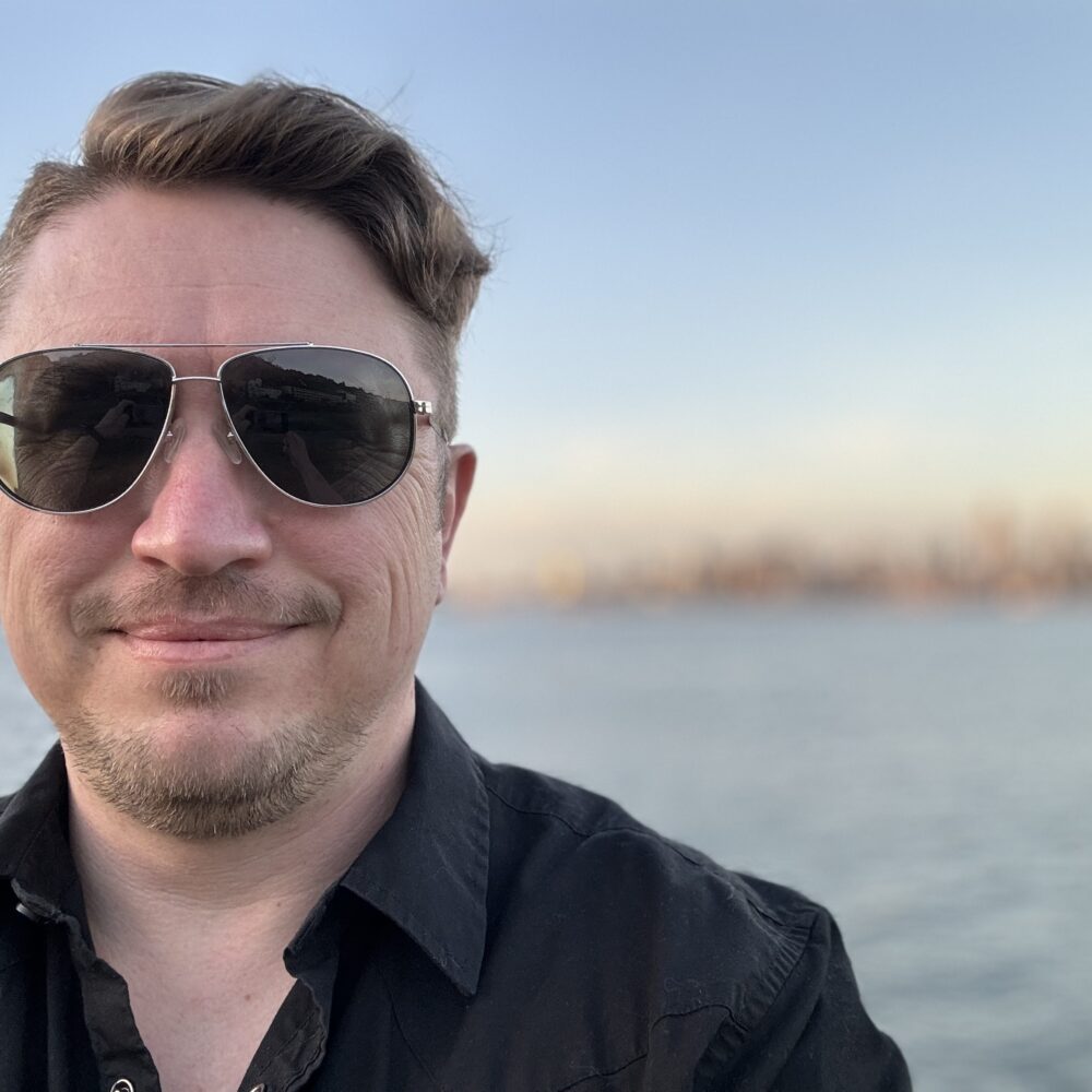 Man in mid-forties with aviator sunglasses stands in front of water with city blurred in the background.